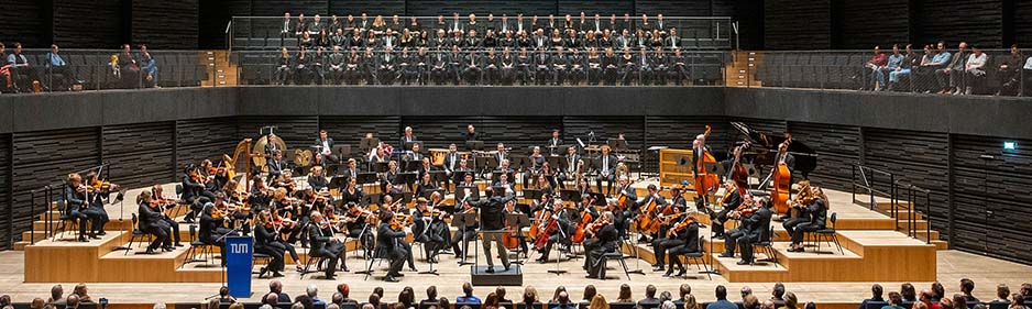 Blick auf die helle Bühne mit Orchstermusikern und Dirigent beim musizieren im Vordergrund. Darüber auf dem Balkon sitzt der TUM Chor. Am unteren Bildrand ist ein Teil des Publikums von hinten zu sehen.